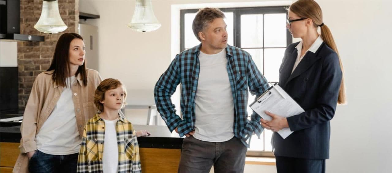 A family of three standing in a modern kitchen, speaking with a real estate agent holding a clipboard.