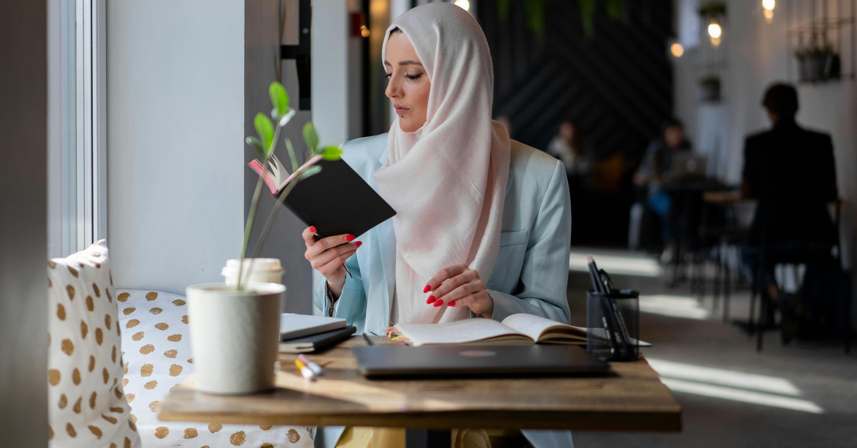 Woman wearing a light-colored hijab and blazer sitting at a cafe table, reading a notebook with a laptop and stationery in front of her