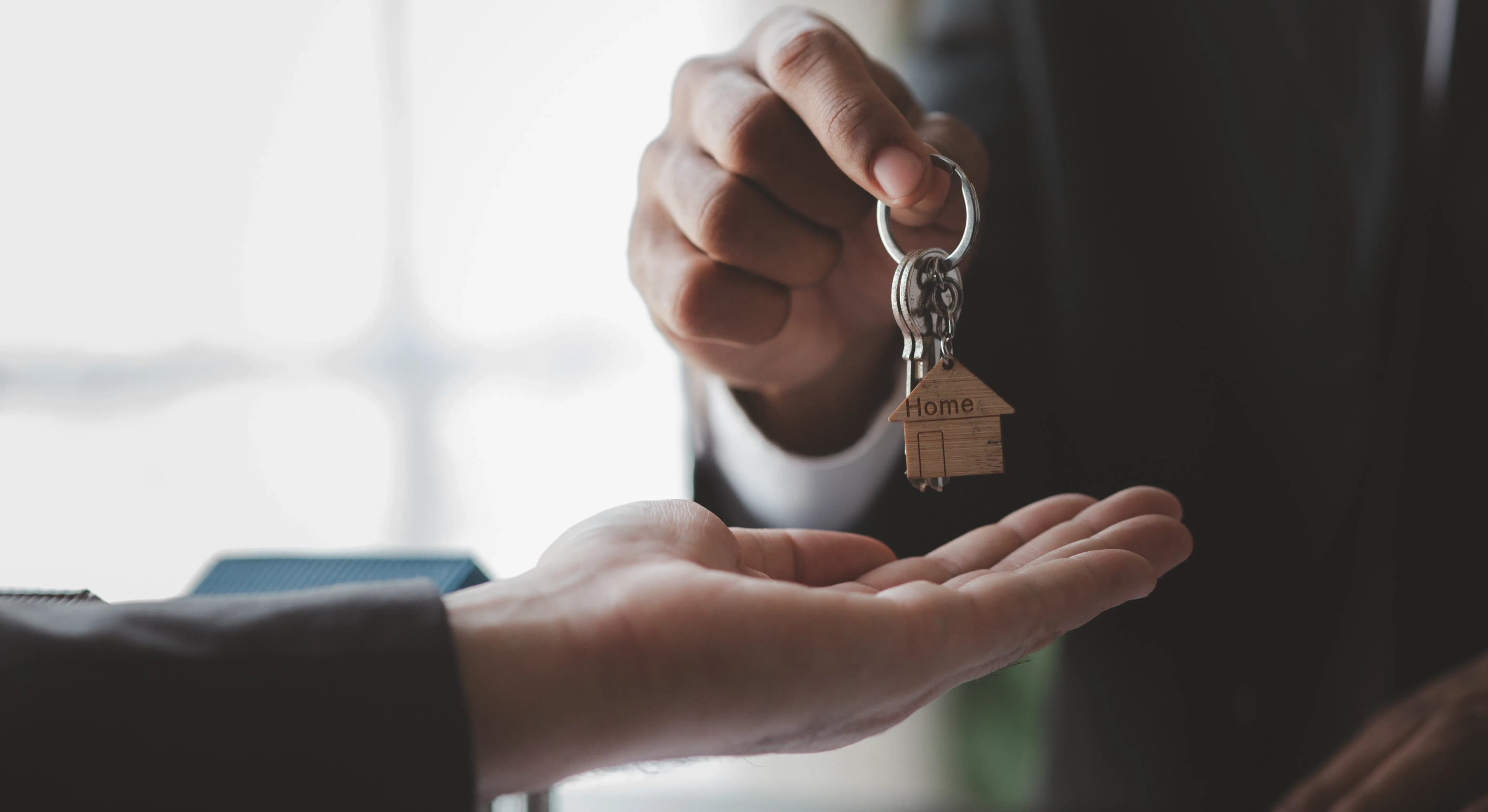 Close-up of a person handing over house keys with a "Home" keychain to another person's outstretched hand indoors, symbolizing real estate transaction or rental agreement.
