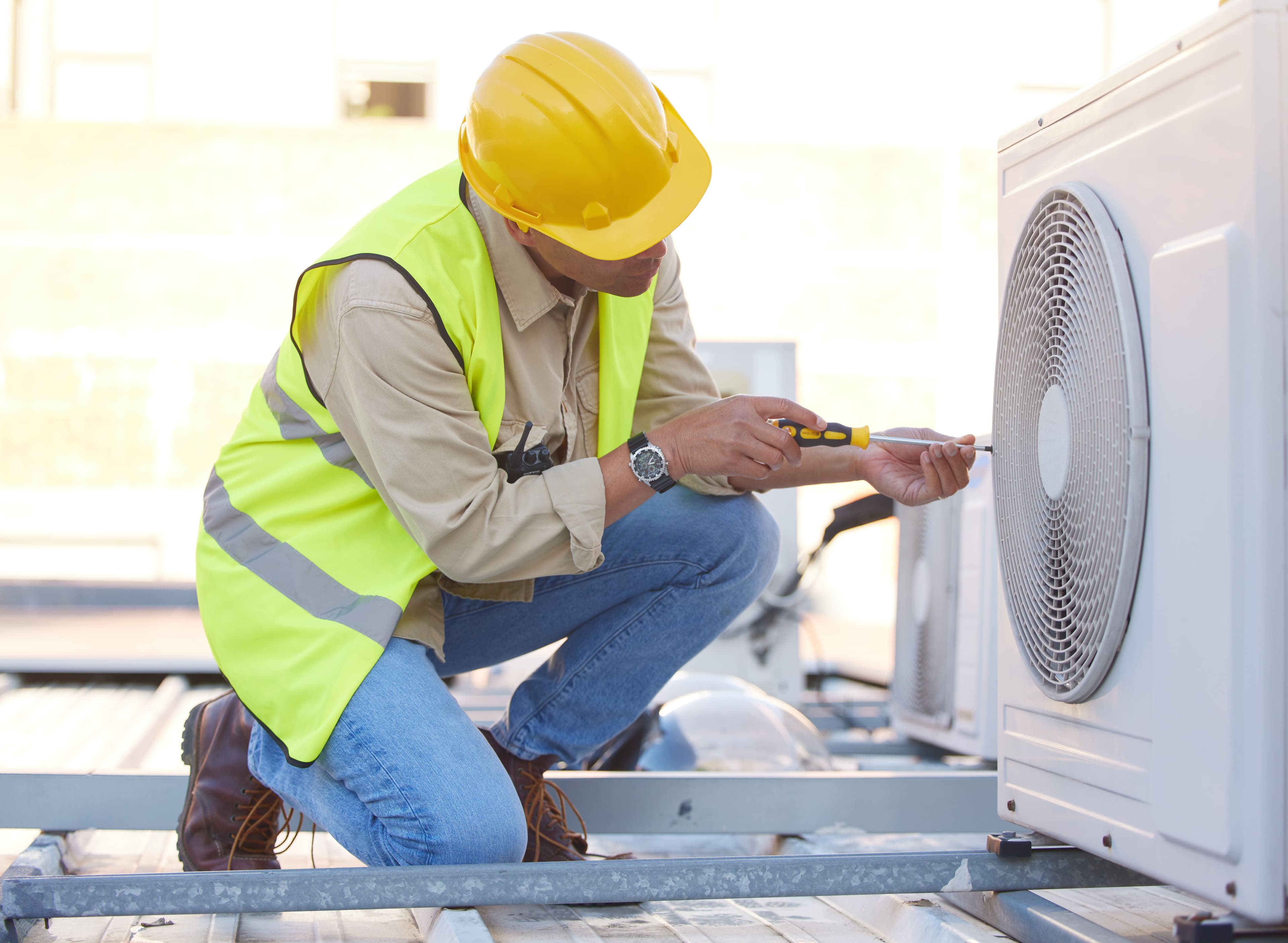 A technician in a yellow safety helmet and vest repairing an outdoor air conditioning unit with a screwdriver.