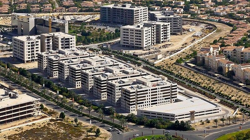 Residential buildings under construction in Dubai Investments Park (DIP) with cranes and palm-lined roads.