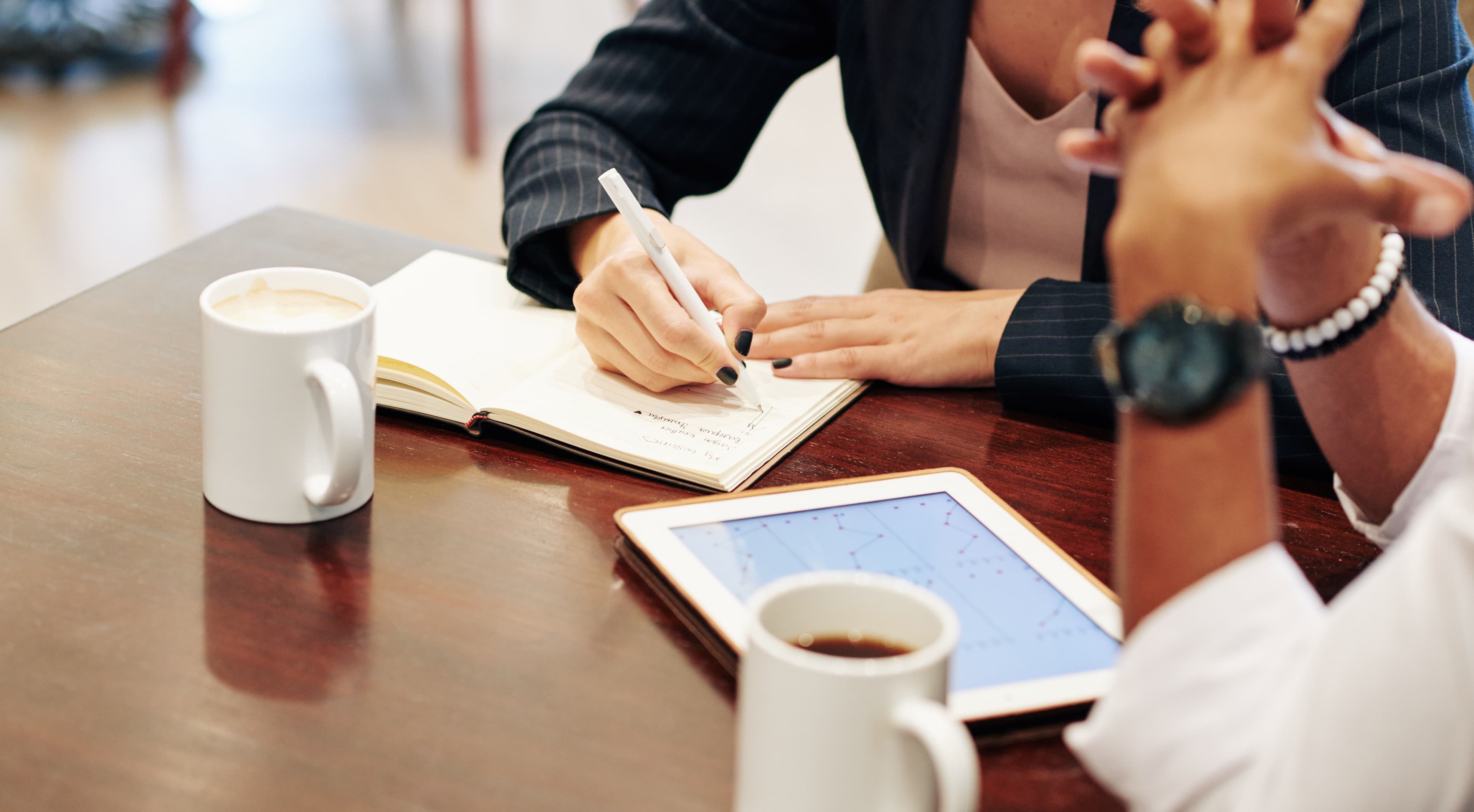 Close-up of a real estate broker writing in a notebook beside a client at a wooden table, with coffee mugs and a tablet showing property market charts, representing a Dubai real estate brokerage consultation.