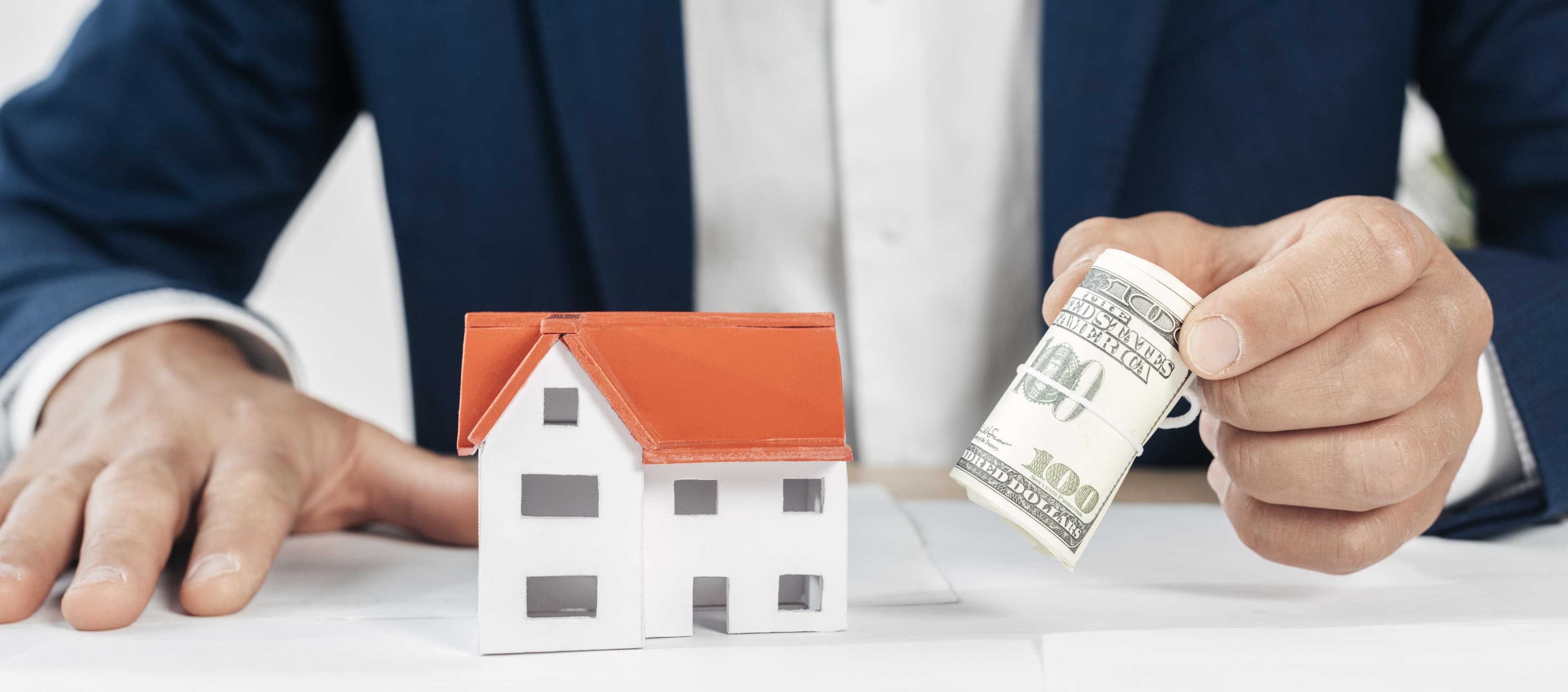 Businessman in a suit holding a roll of US dollar bills next to a small model house on a white desk.