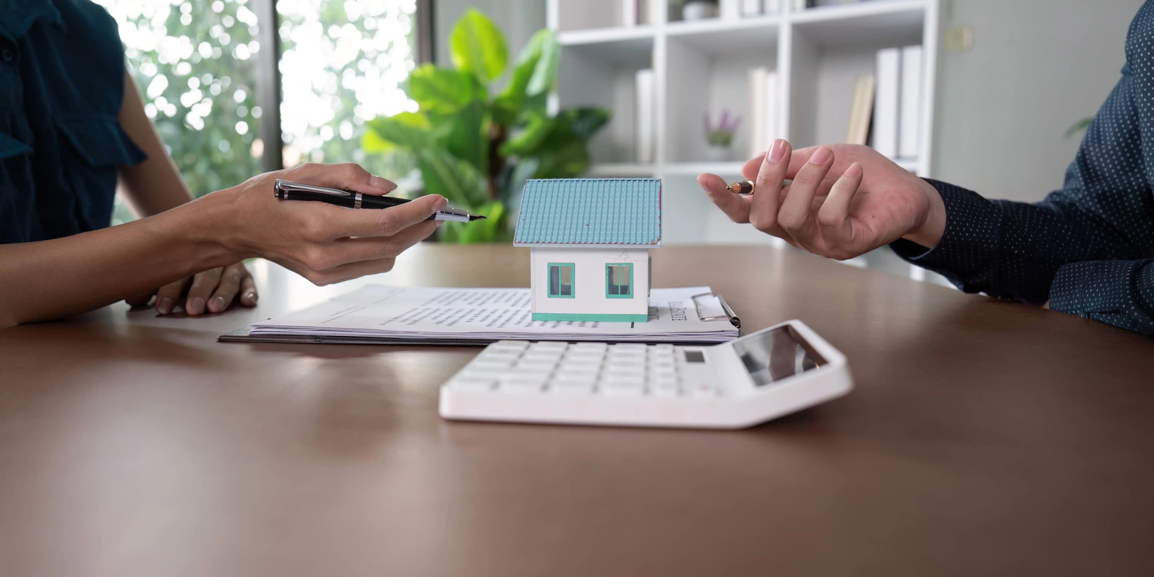 Two individuals at a desk, one holding a pen, with a small model house, papers, and a calculator in view. The setting suggests a discussion about real estate or financial planning.
