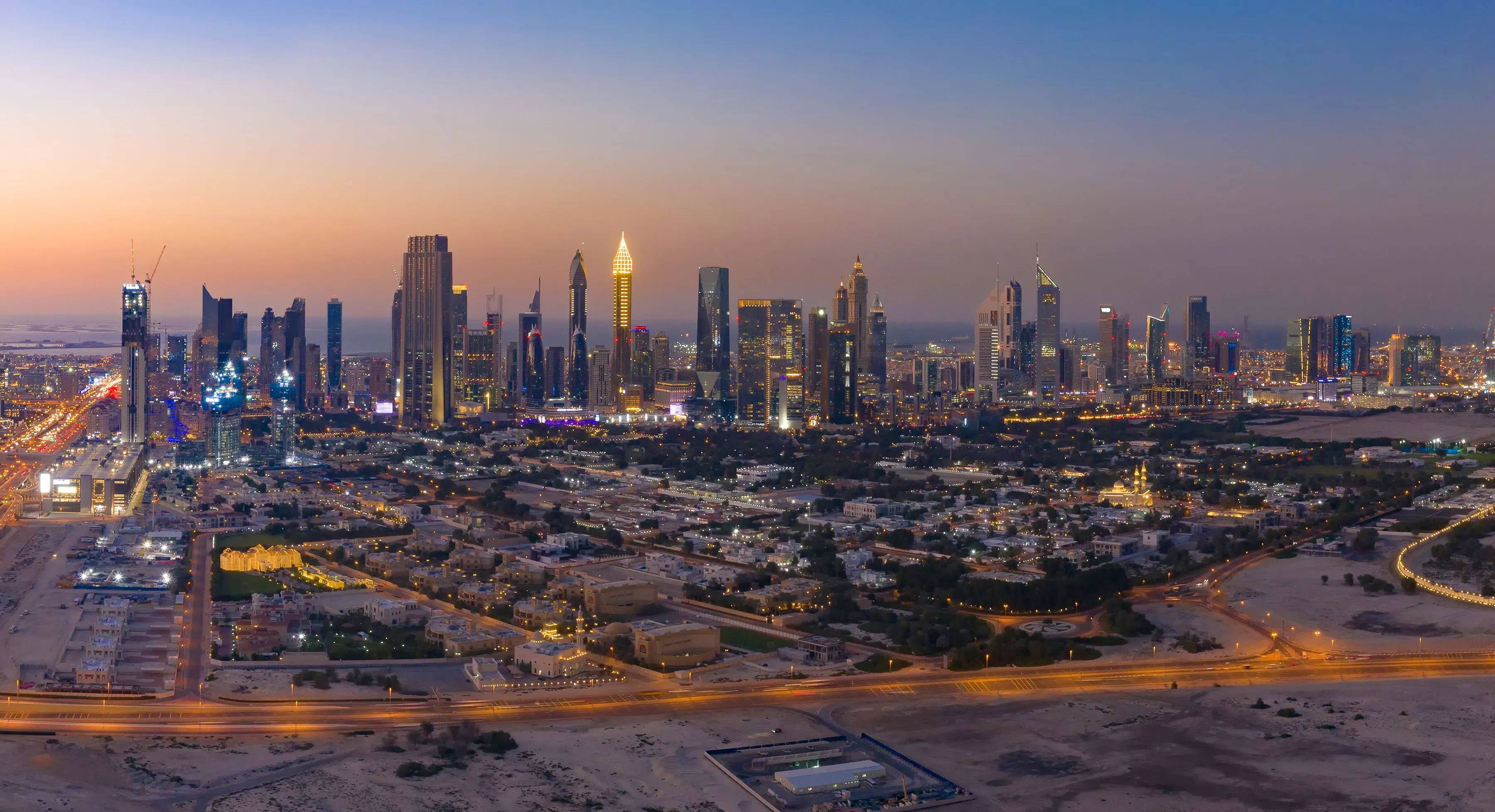 An aerial shot of a city at dusk, highlighting a blend of skyscrapers and residential neighborhoods, with glowing lights illuminating the streets and buildings.