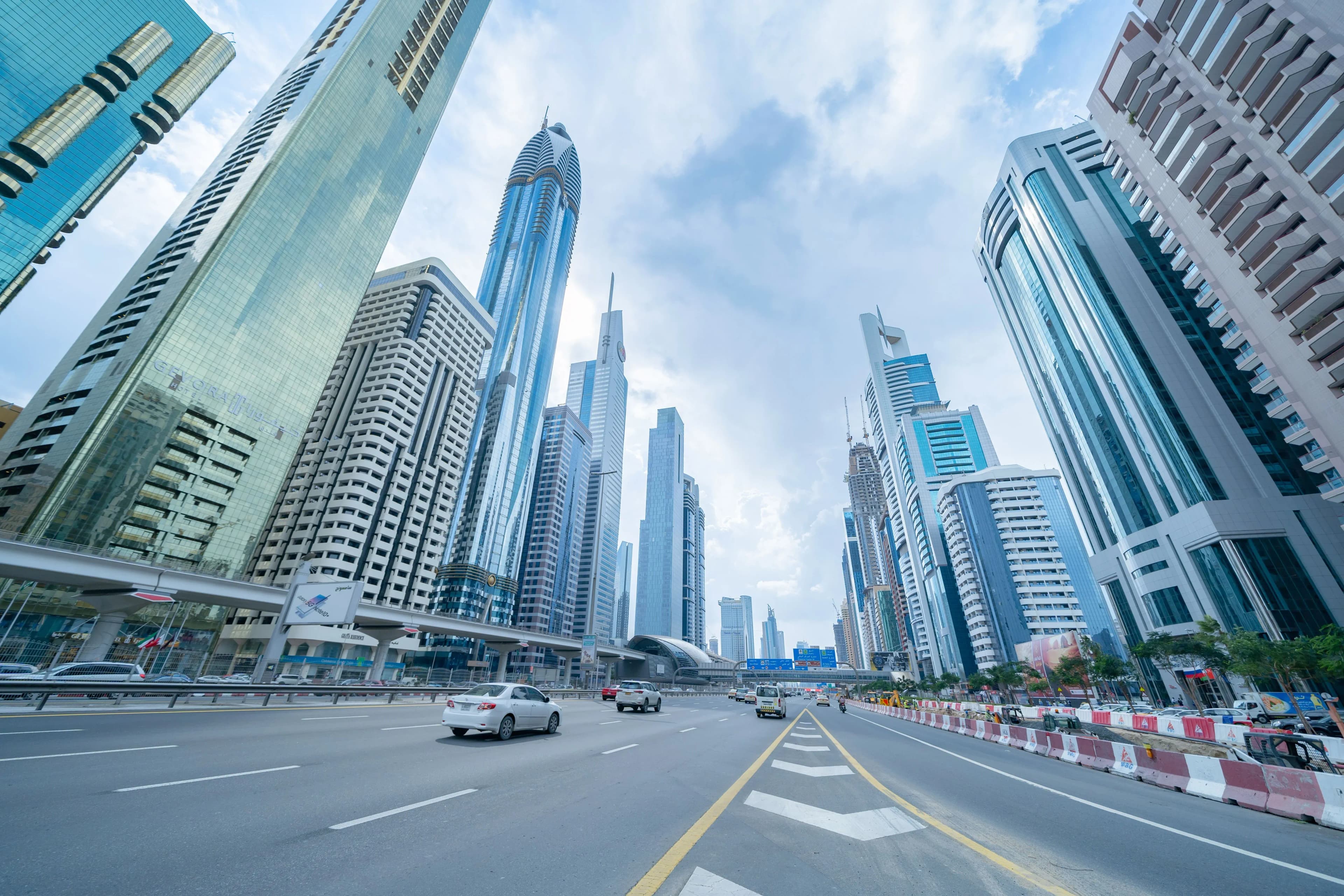A low-angle street-level view looking up at the towering glass skyscrapers lining Sheikh Zayed Road, with cars driving on the wide highway under a partly cloudy sky.