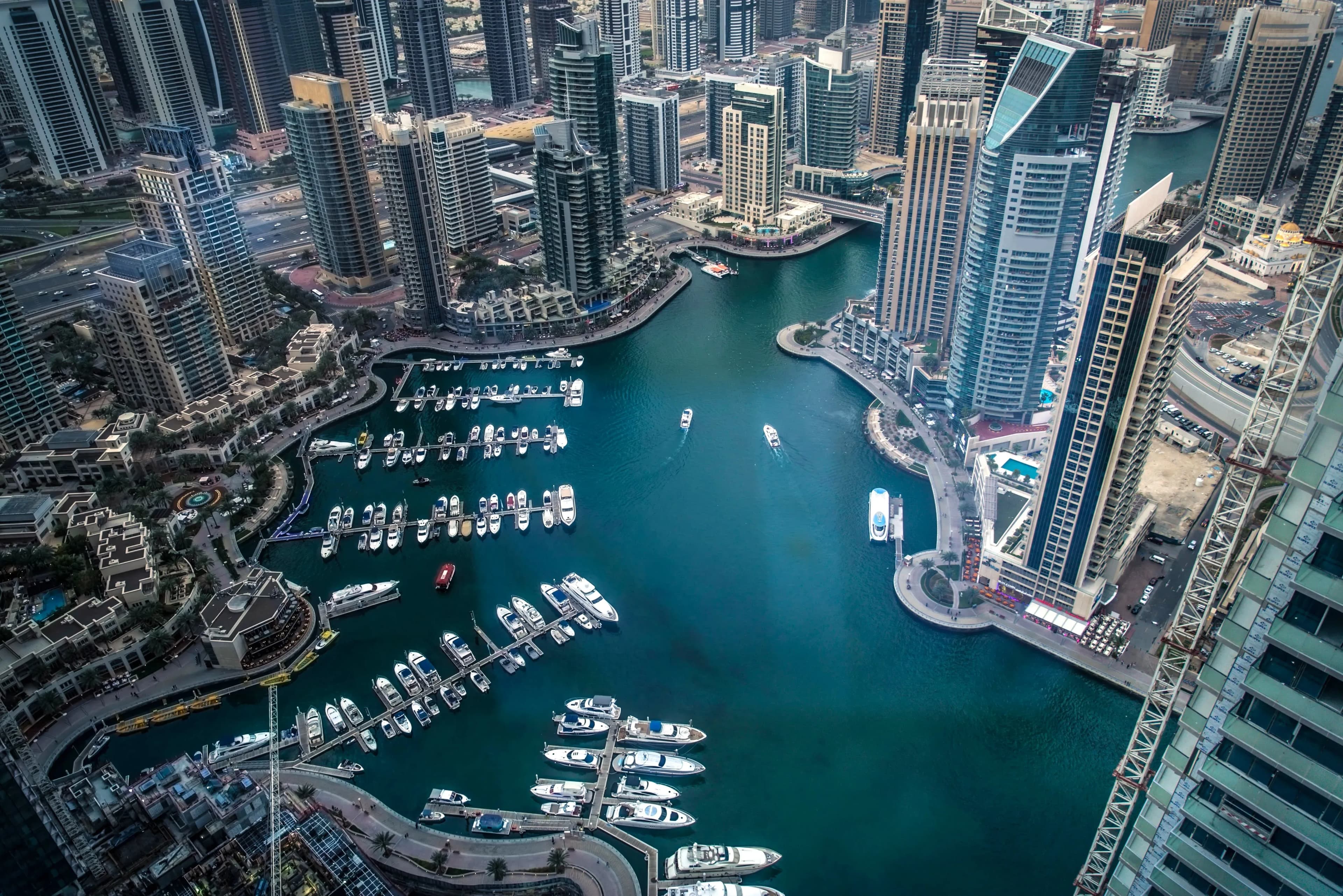 Aerial view of Dubai Marina with clusters of tall buildings surrounding turquoise water, marinas full of yachts, and boats cruising through the channel.