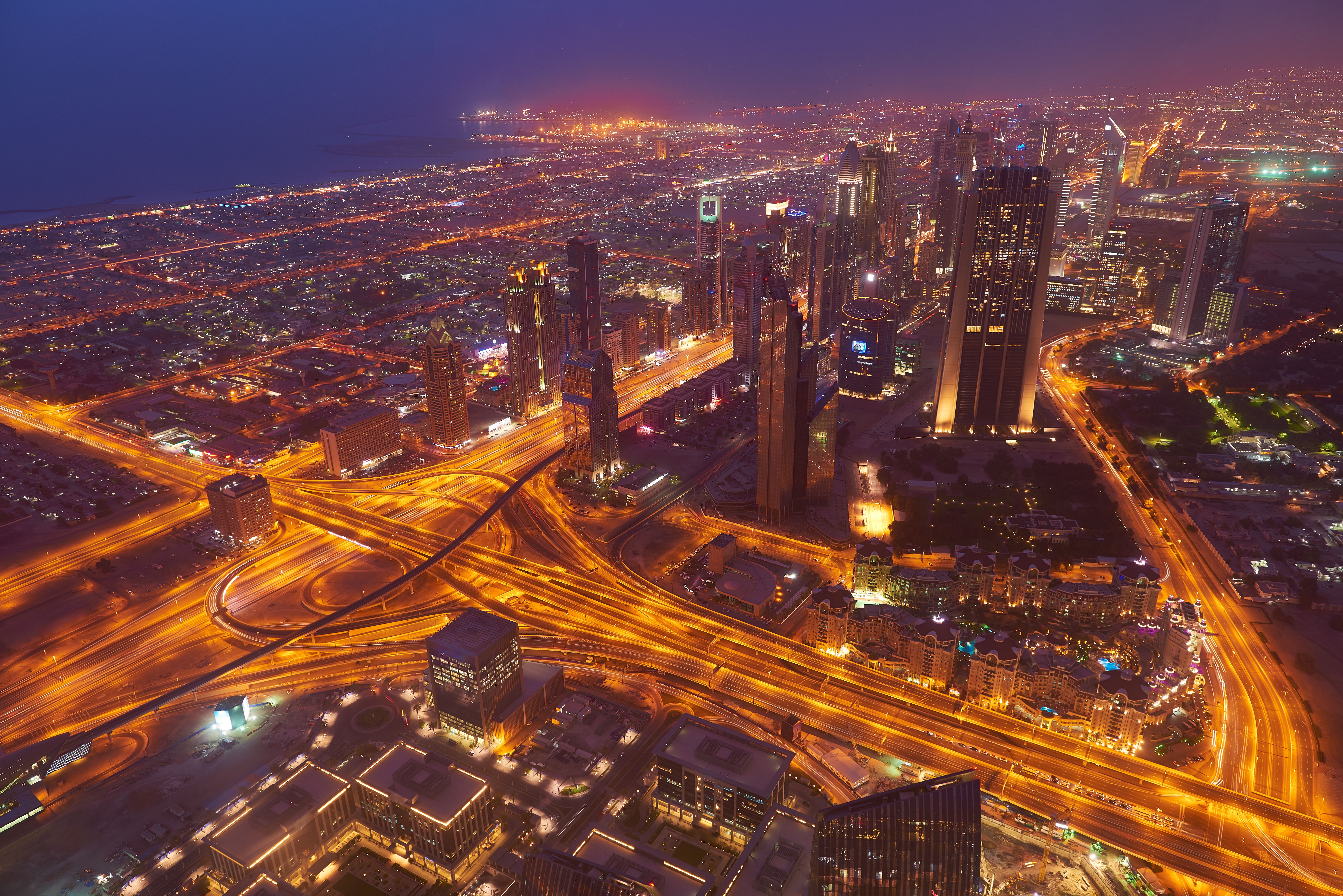 An aerial night view of a brightly illuminated city with tall skyscrapers and complex highways, showing Dubai’s iconic skyline.