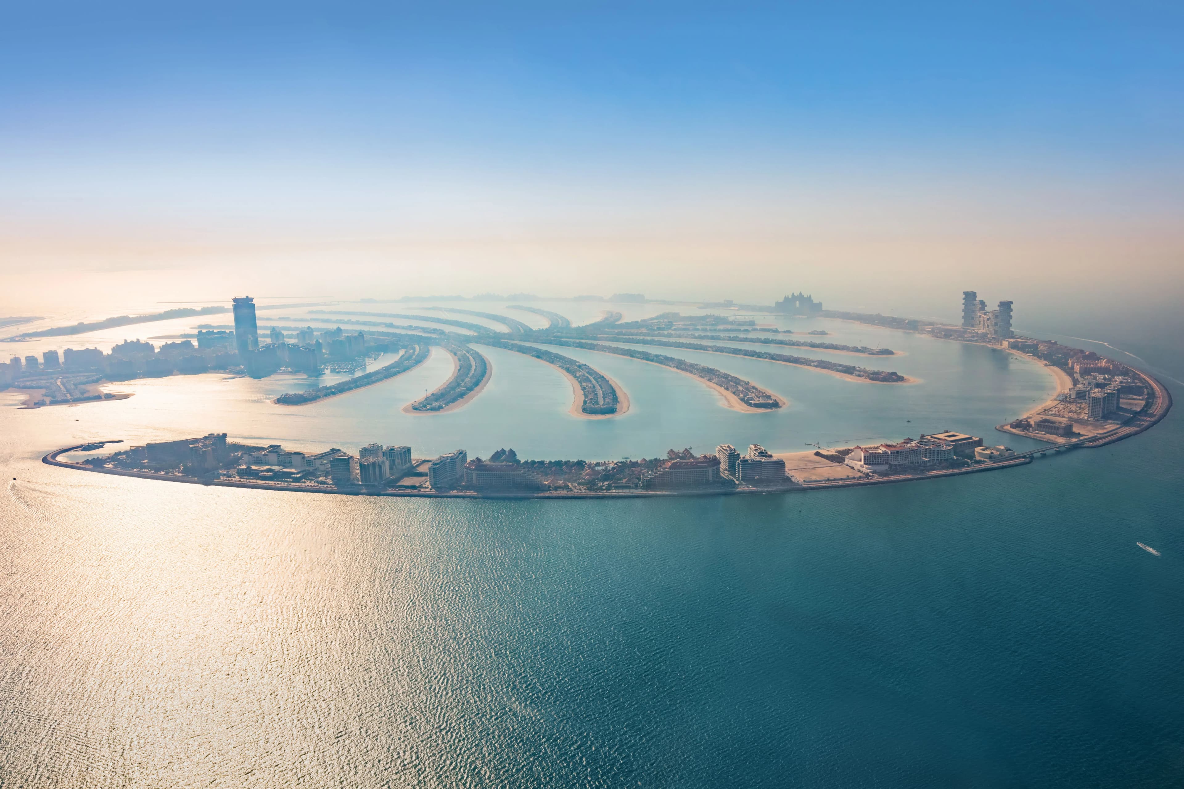 A sweeping aerial shot of the entire Palm Jumeirah island, highlighting its iconic palm tree shape, fronds lined with villas, and the surrounding blue sea under a hazy sky.