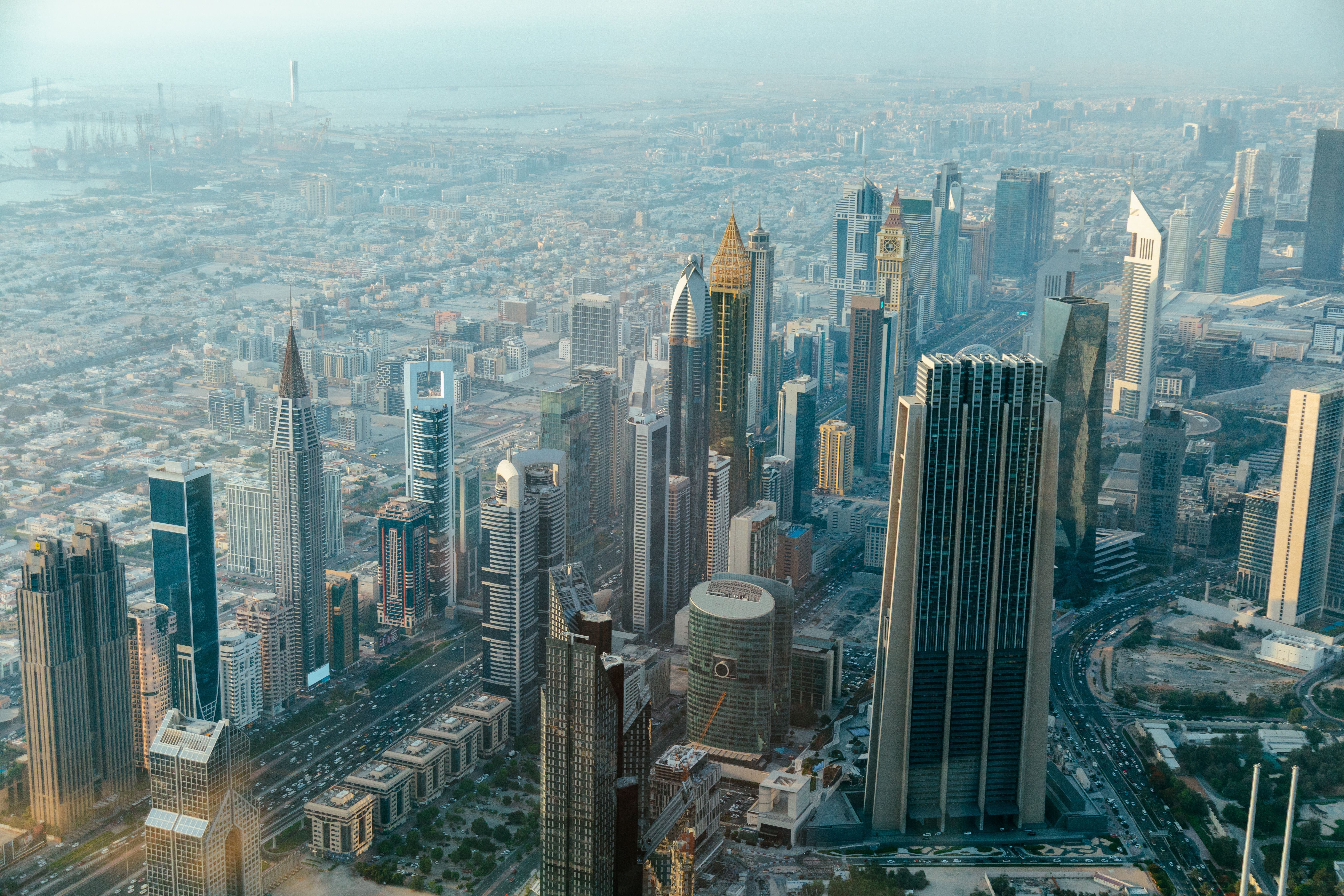 High-angle photo showing a dense cluster of modern skyscrapers in downtown Dubai, with roads and buildings stretching into the distance.