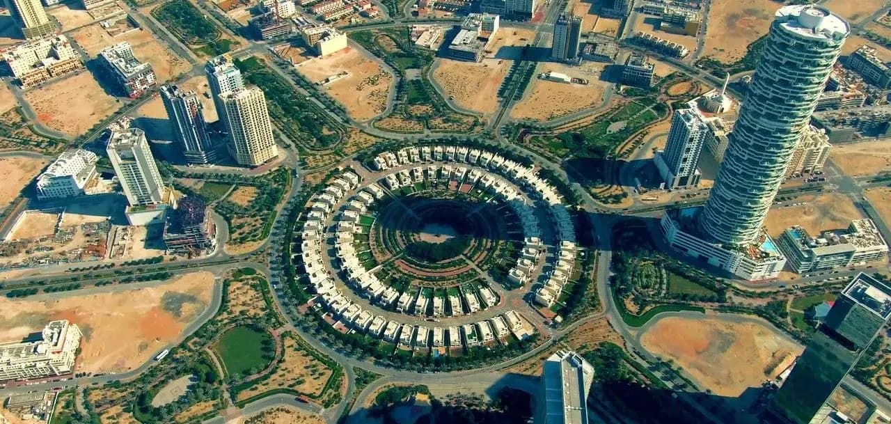 Aerial photograph of Jumeirah Village Circle in Dubai, showing concentric rings of white villas with surrounding roads and high-rise towers in a desert urban setting.