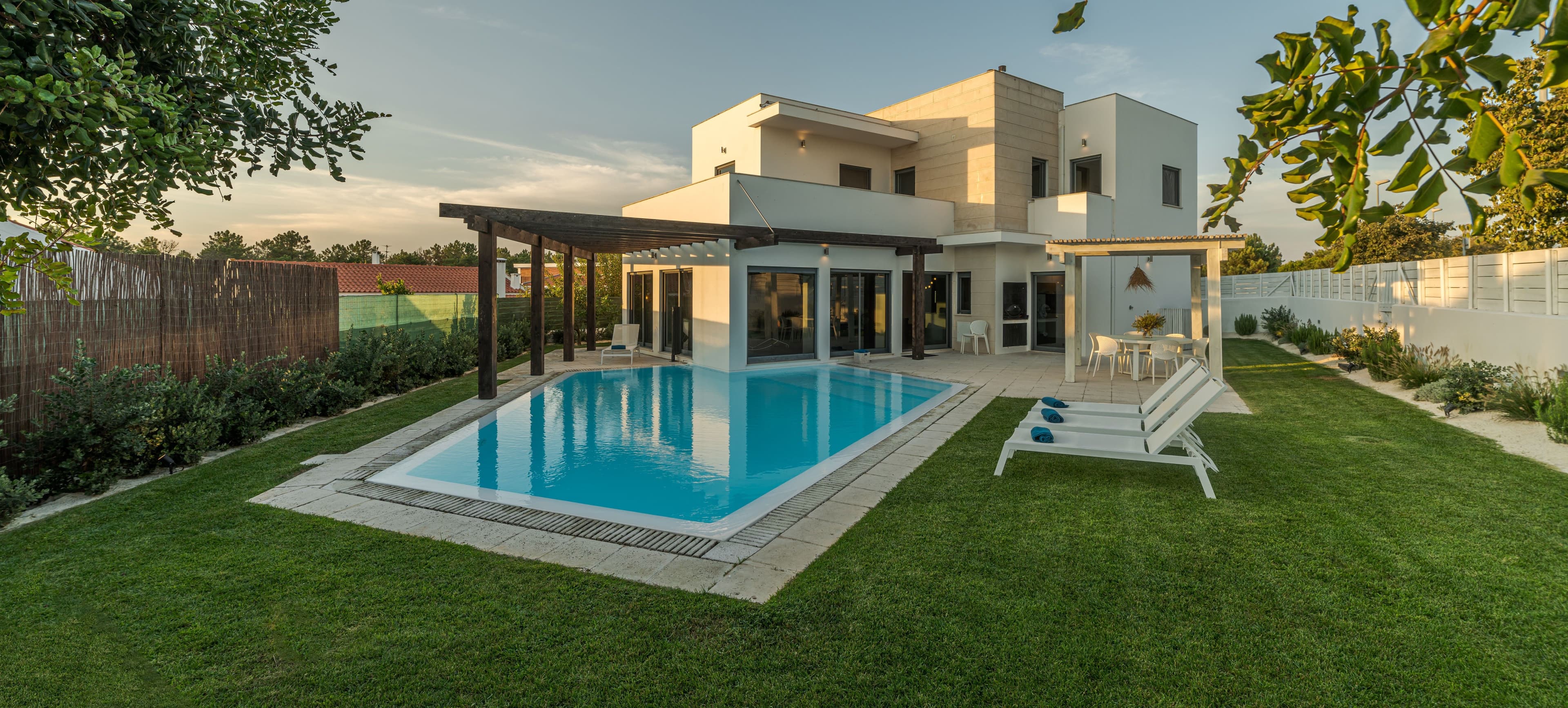 Contemporary white villa featuring a spacious backyard with a rectangular swimming pool, green lawn, sun loungers, and a shaded outdoor dining area, photographed in the evening light.