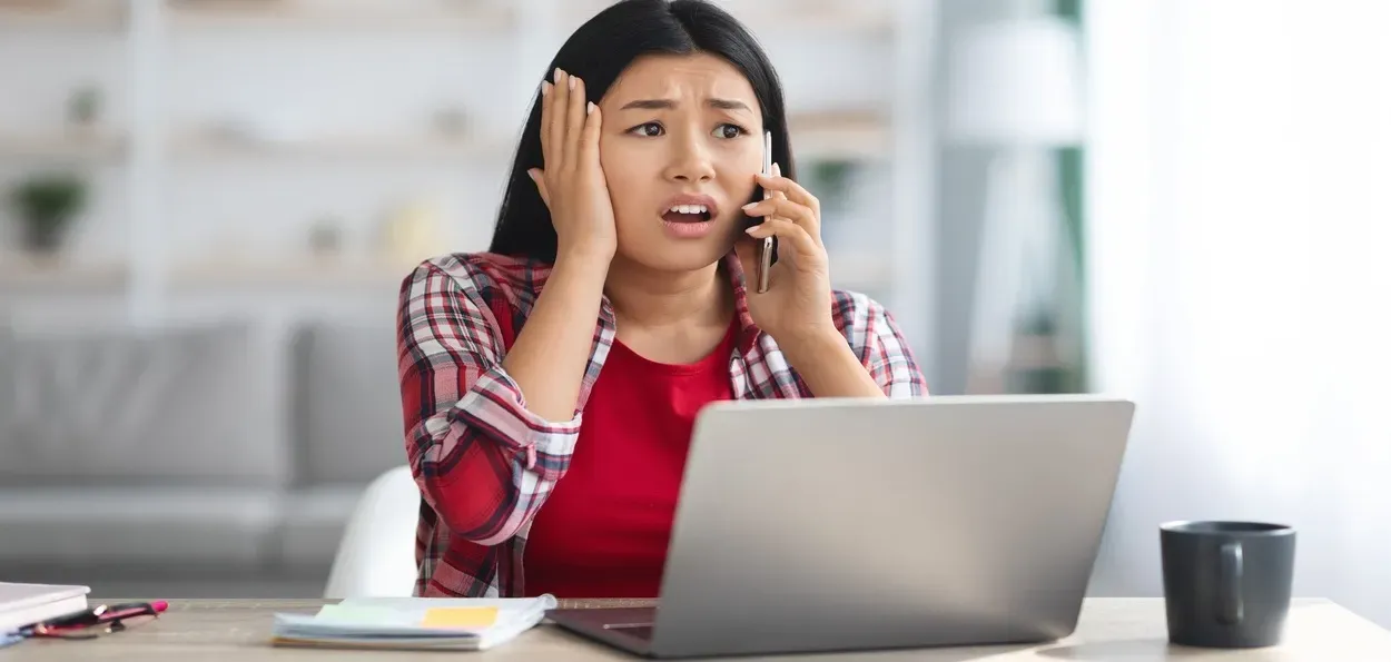 A woman in a red shirt sits at a desk with a laptop, holding her head in one hand and a phone in the other, looking worried.
