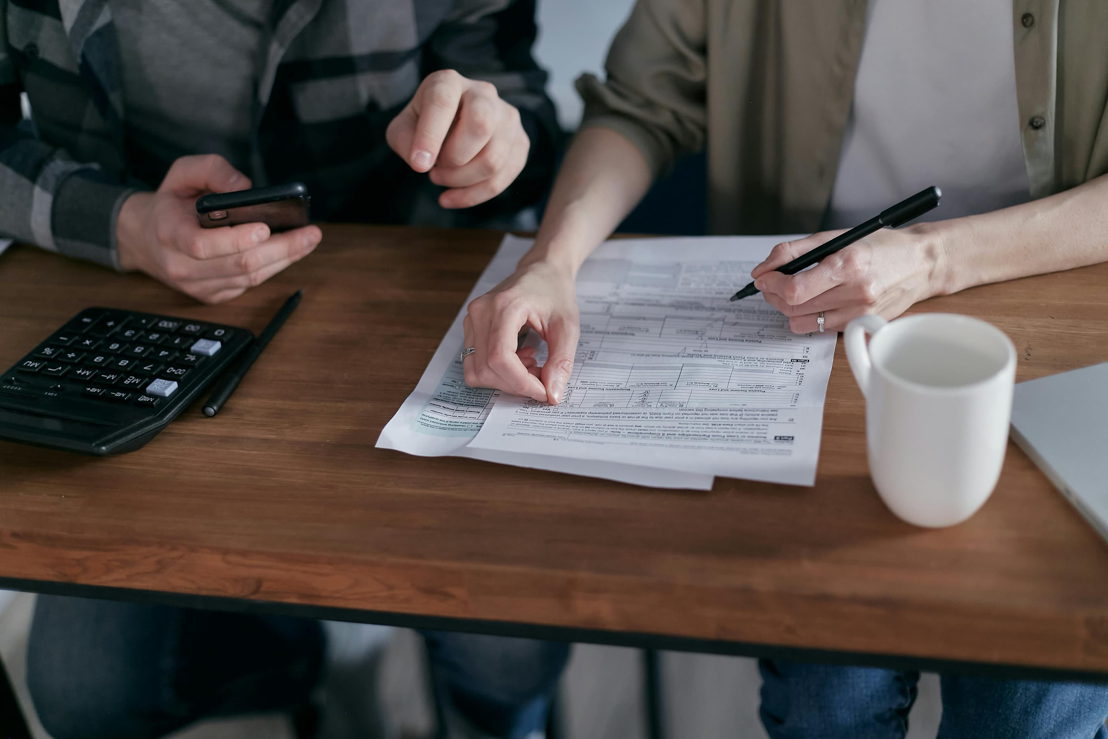 Two people sitting at a table, filling out forms with a pen, calculator, smartphone, and coffee mug nearby, suggesting budgeting or handling important documents.