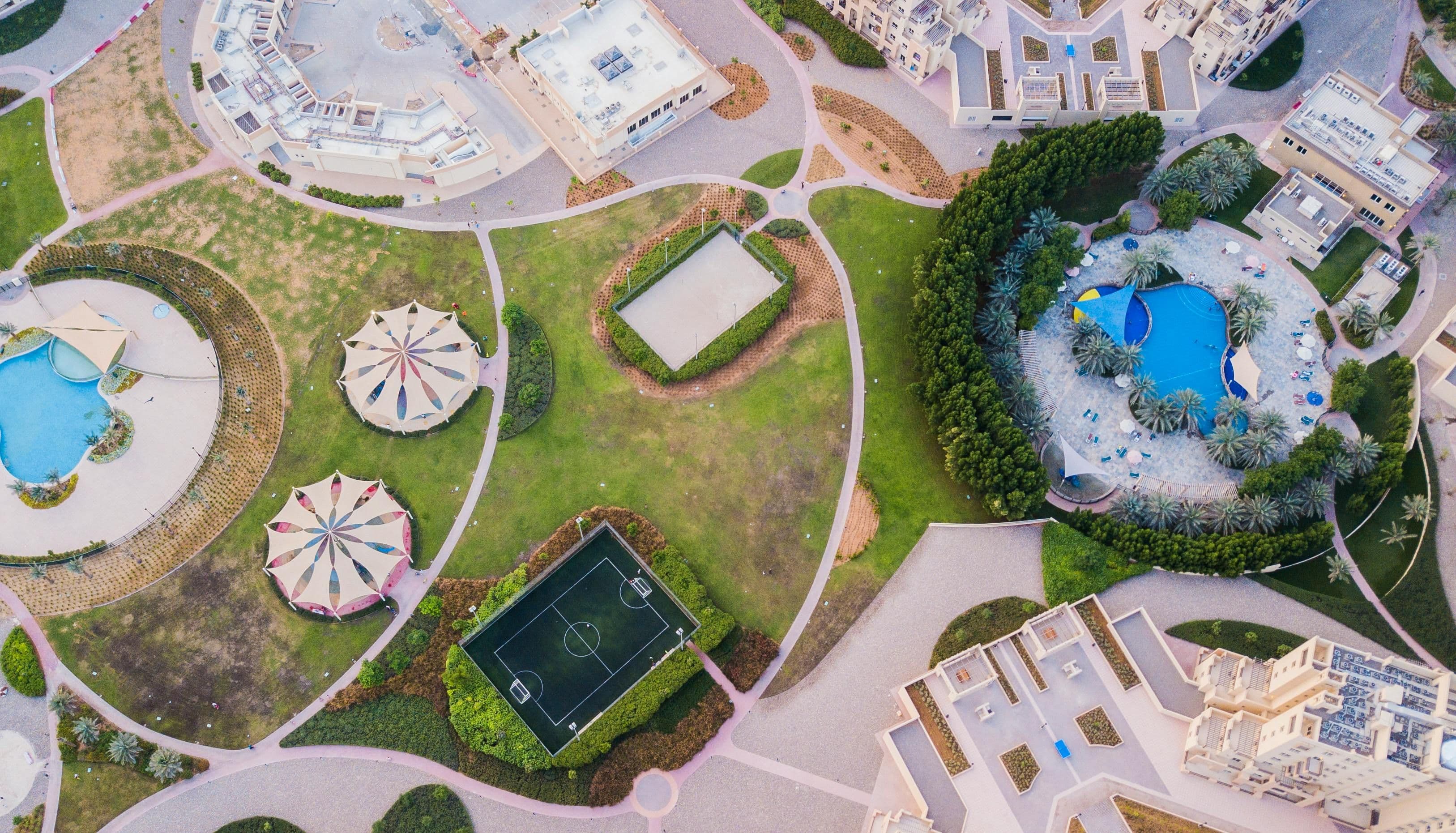 Aerial view of a landscaped residential park with walking paths, sports courts, playground canopies, and a swimming pool surrounded by trees and buildings.