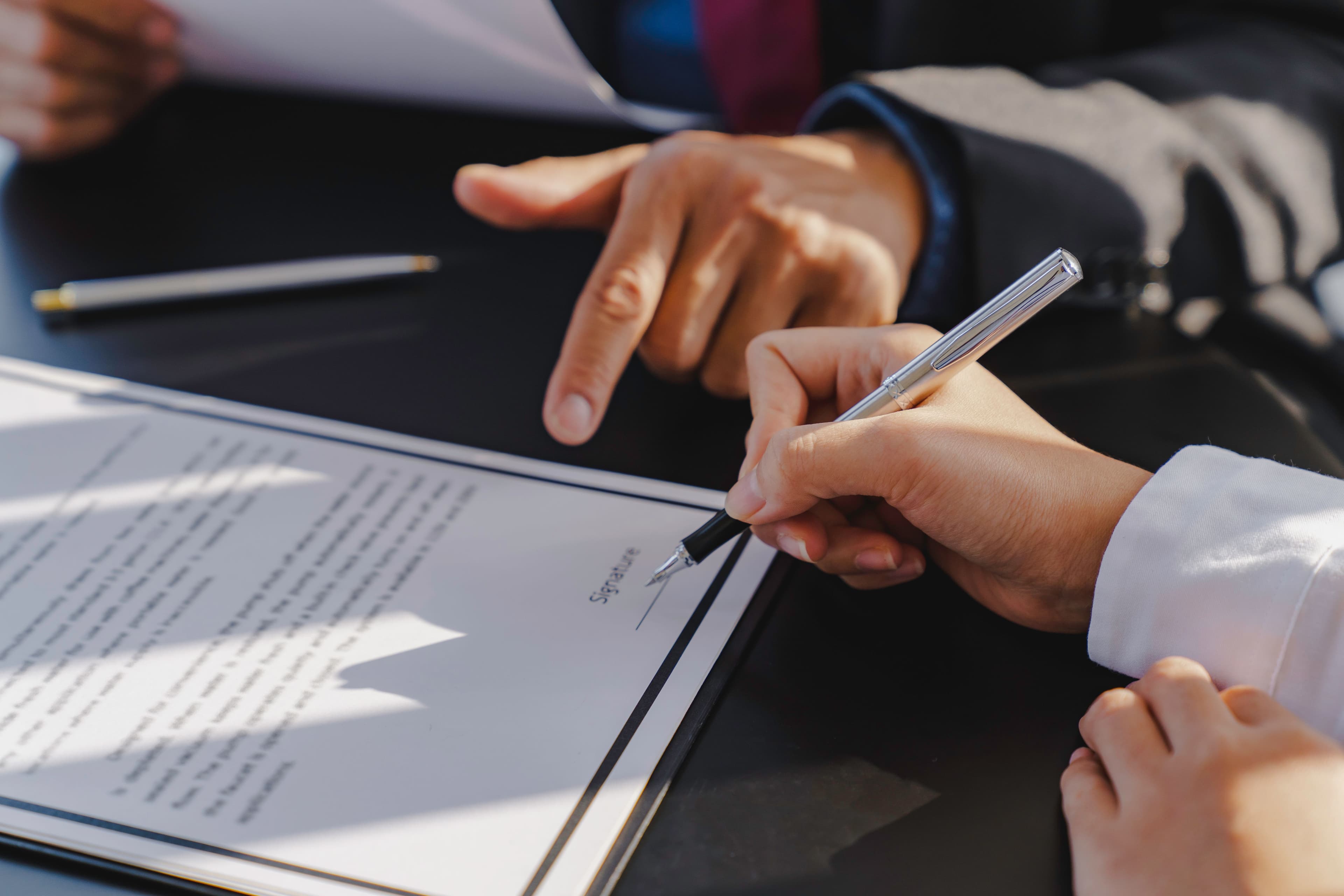 Close-up of a person signing a real estate contract with a pen while a professional points to the document on a desk.