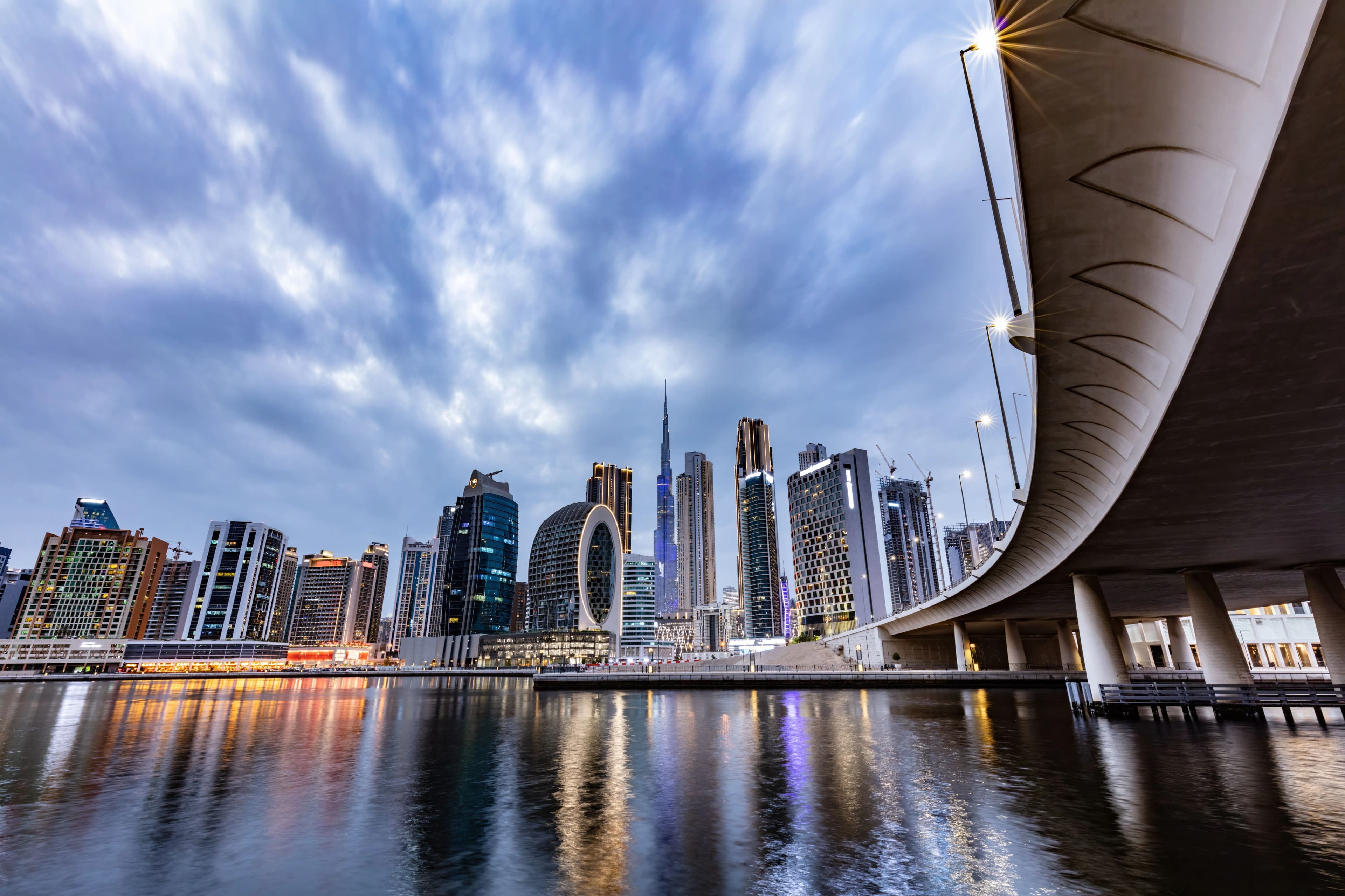 Waterfront view of Dubai Downtown skyline at dusk with the Burj Khalifa rising among modern skyscrapers and a curving bridge in the foreground.