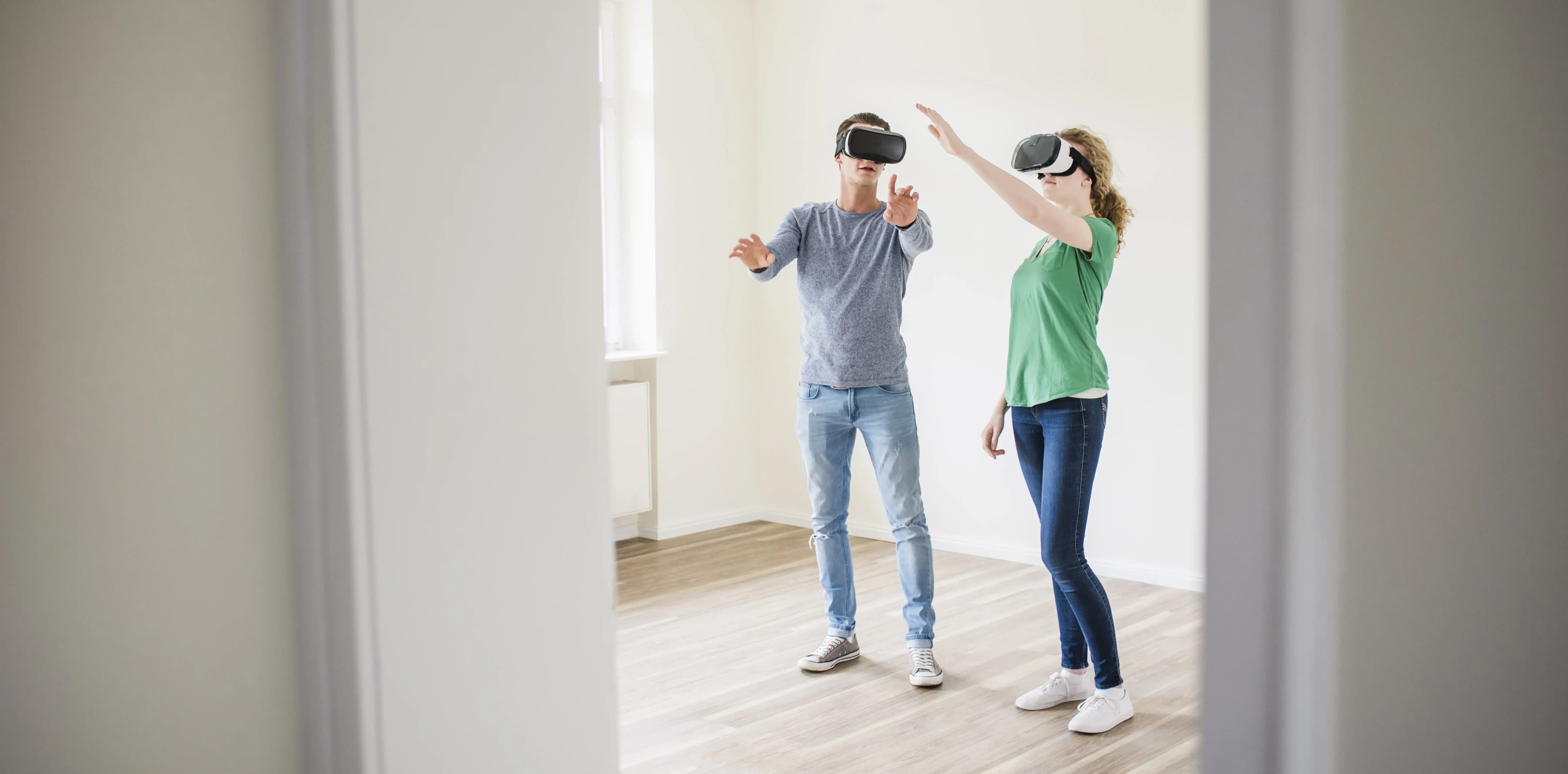Two people standing in an empty, light-filled apartment wearing VR headsets, gesturing as they interact with a virtual environment to visualize space and design options.